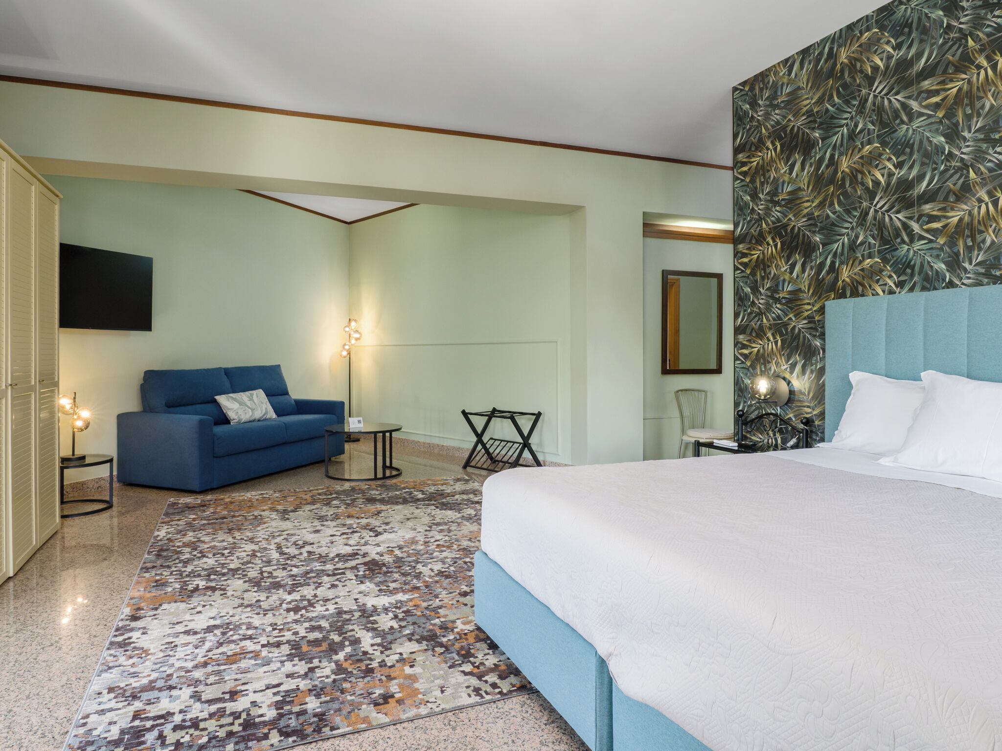 Habitación de hotel con una cama, un sofá azul, una mesita, una silla, un televisor y un papel tapiz estampado.