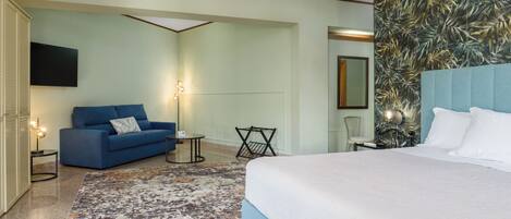 A hotel room with a bed, a blue sofa, a small table, a chair, a TV, and a patterned wall.
