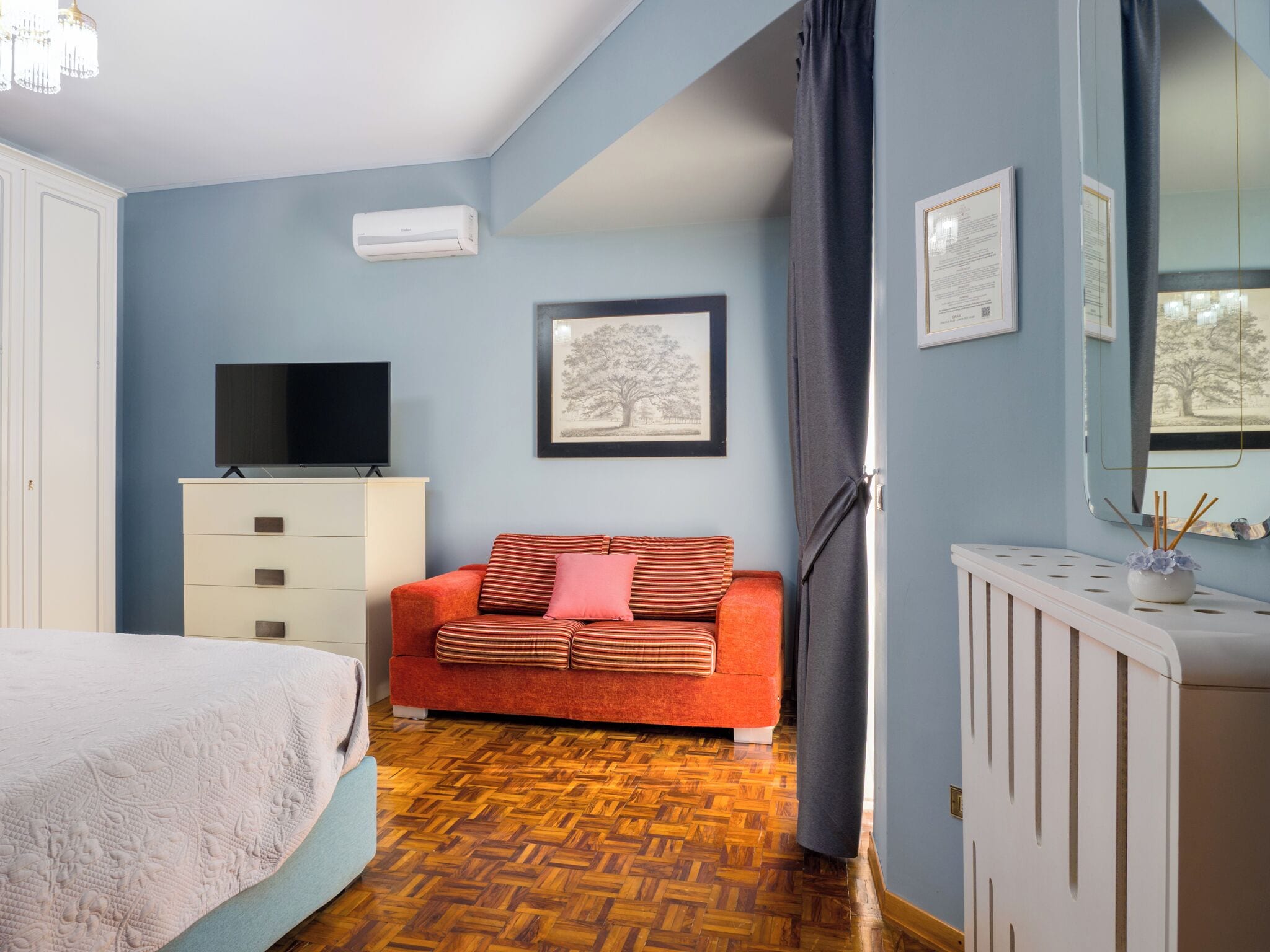 A bedroom with a bed, a television on a dresser, a red sofa, a framed picture on the wall, and a radiator.