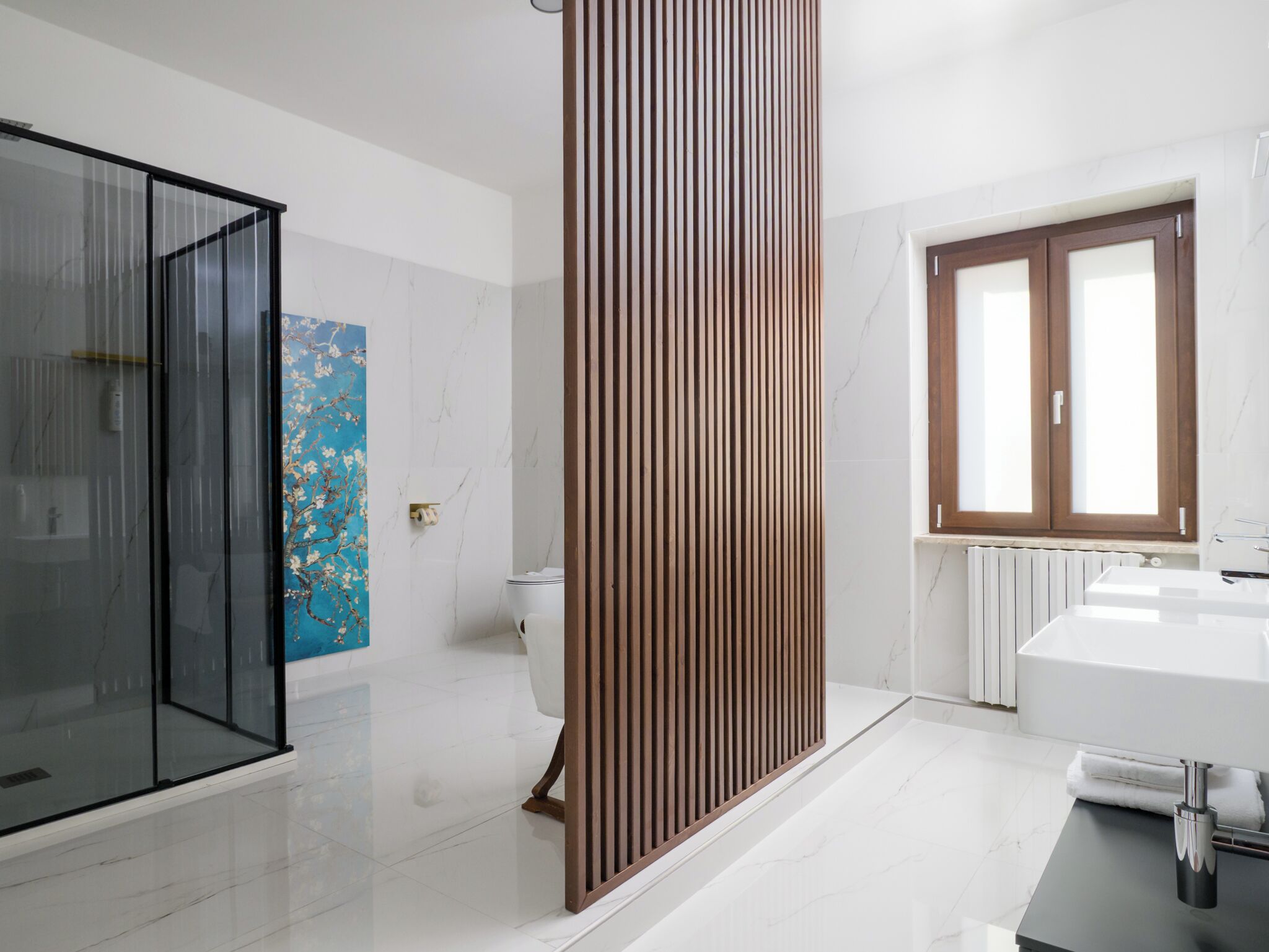 A modern bathroom with a glass shower enclosure, a wooden privacy screen, a marble floor, a white sink, and a window with wooden frames.