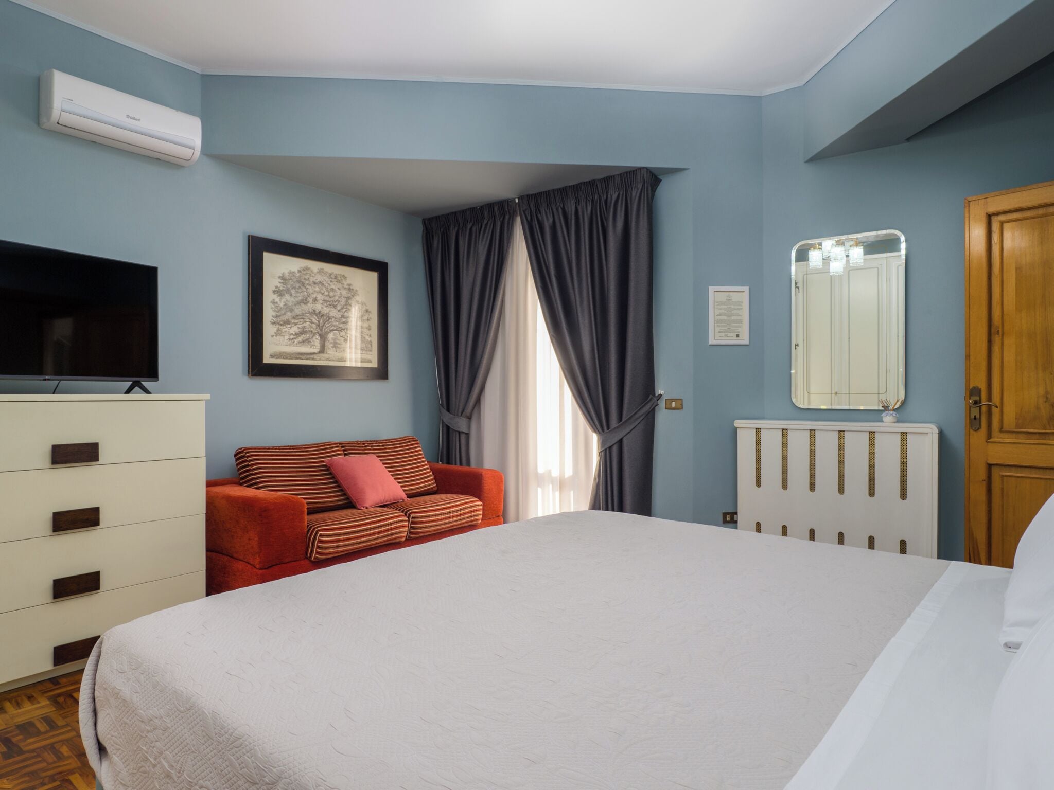 A bedroom with a bed, a dresser, a television, a framed picture, a window with curtains, and a radiator.