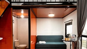 A compact room with a sofa, a small table with items, a window with curtains, and a visible toilet. - Krincing Hills Cabin (Kebonlegi)