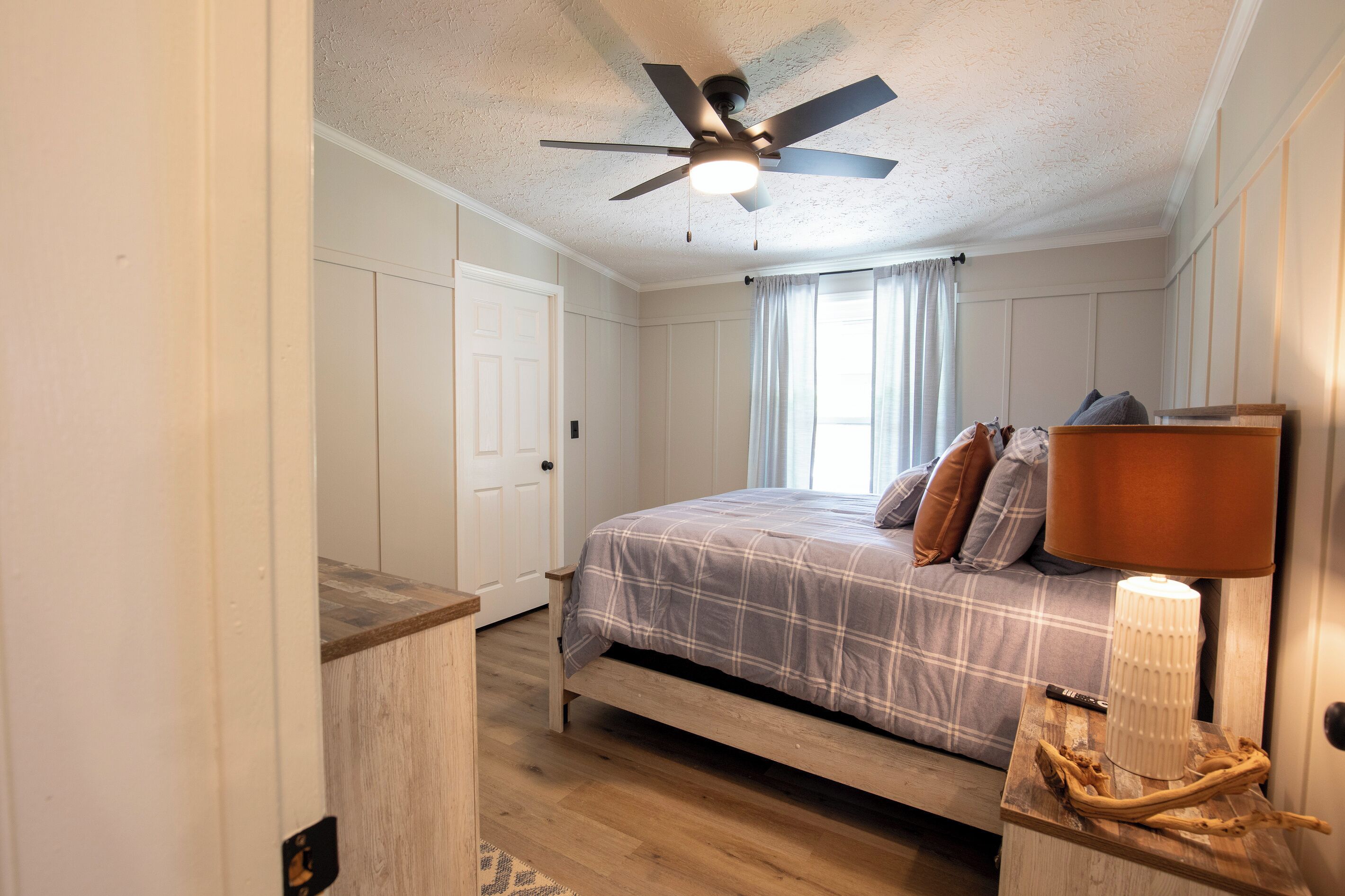 Un dormitorio con cama, ventilador de techo, lámpara de noche y ventana con cortinas.
