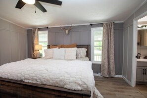 A bedroom with a bed, a ceiling fan, a window with curtains, and a kitchen area visible through an open door.