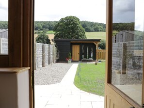 A view through a doorway to a small building with a wooden door, a paved path leading to it, and a grassy area with a fence. - The Old Farmhouse, Westhope (Craven Arms)