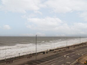 Eine Küstenstraße mit Strand rechts, mit einer Reihe Bänken und einem Zaun entlang der Uferpromenade.