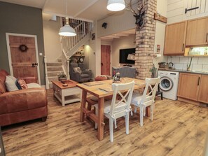A spacious living area with a wooden dining table, white chairs, a sofa, a staircase, and a kitchen area with wooden cabinets and a washing machine.