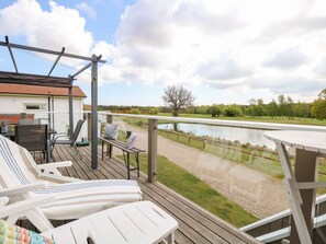 A deck with white lounge chairs, a table, and a view of a lake and park. - Links View Retreat in Fritton (Great Yarmouth)