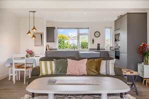 A modern kitchen with a large window, a dining area with a white table and chairs, and a living area with a sofa and cushions. - Wide Horizons, Coverack (Helston)