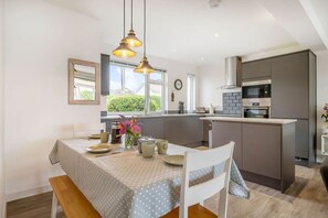 A modern kitchen with a dining area, featuring a central island, pendant lighting, and a view of greenery outside. - Wide Horizons, Coverack (Helston)