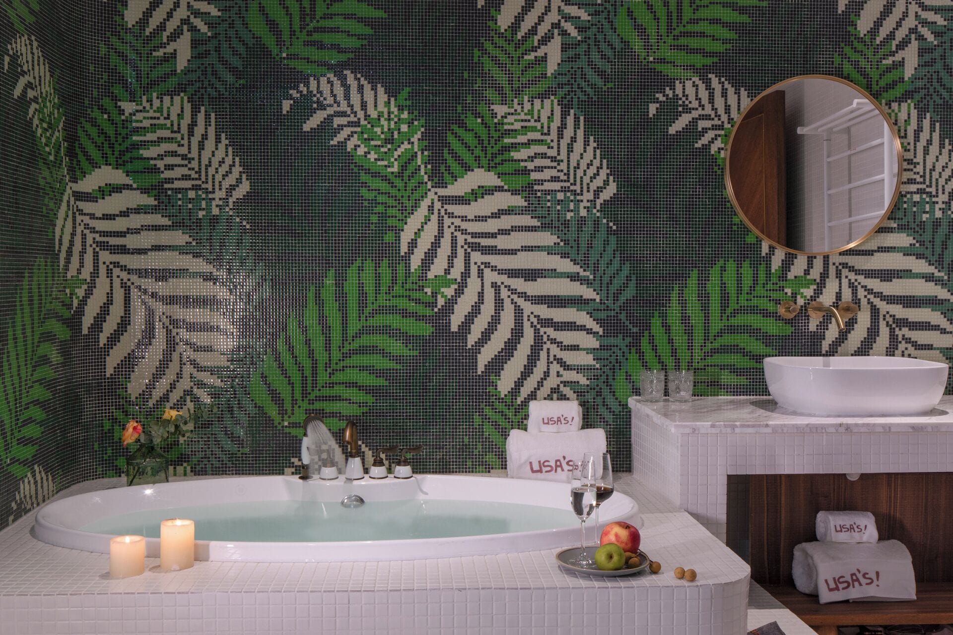 A bathroom with a large bathtub, a sink, and a mirror. There are towels with 'USA's' written on them, a plate with fruit, and candles.