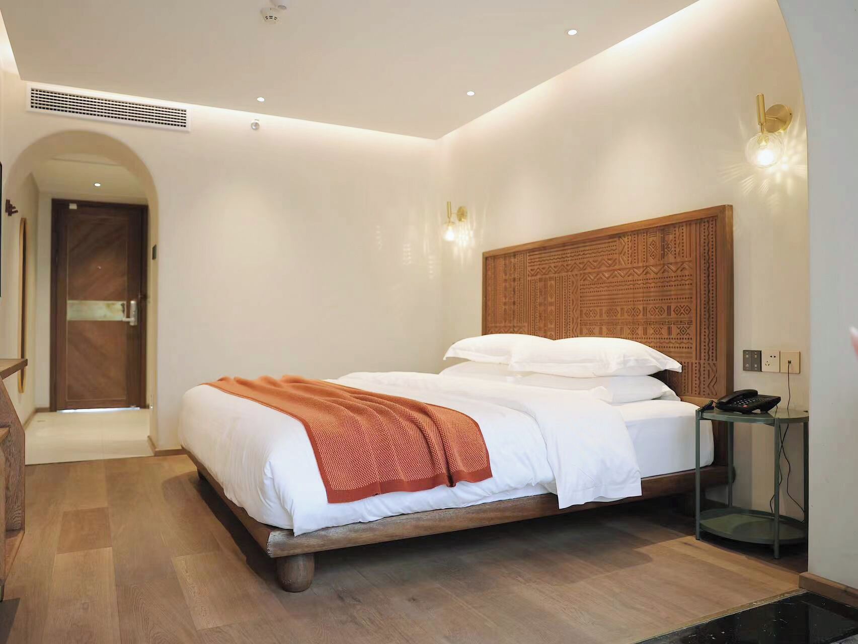 Une chambre d’hôtel moderne avec un lit en bois, une tête de lit intégrée, une table de chevet avec un téléphone et une porte donnant sur un couloir.