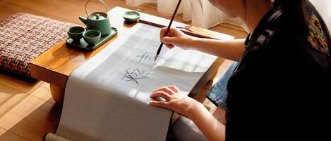 A person sitting on a wooden floor, writing on a large piece of paper with a brush. There is a teapot and cups on a tray beside them.
