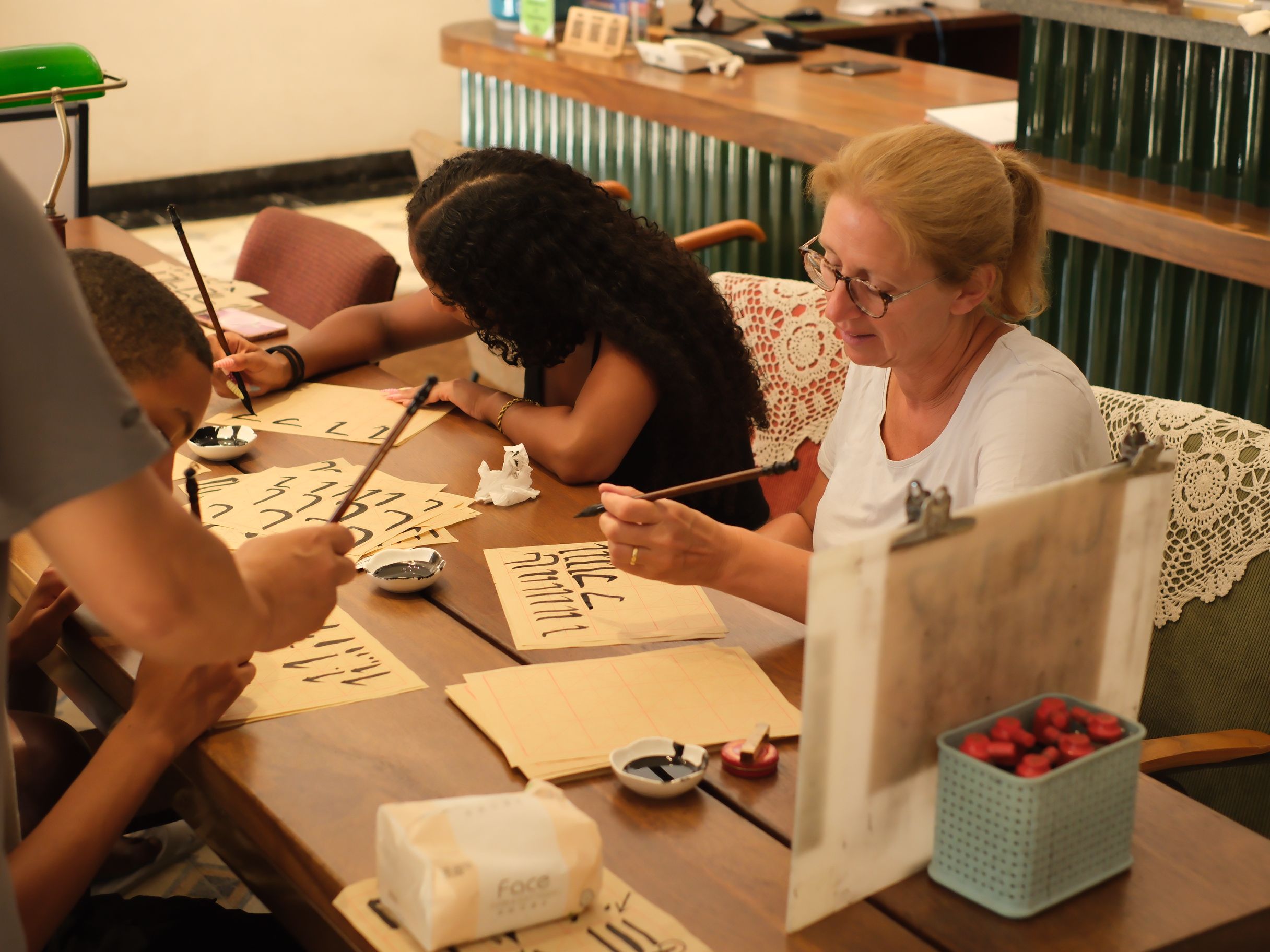 A group of people engaged in a calligraphy activity at a wooden table with paper, brushes, and ink bowls.