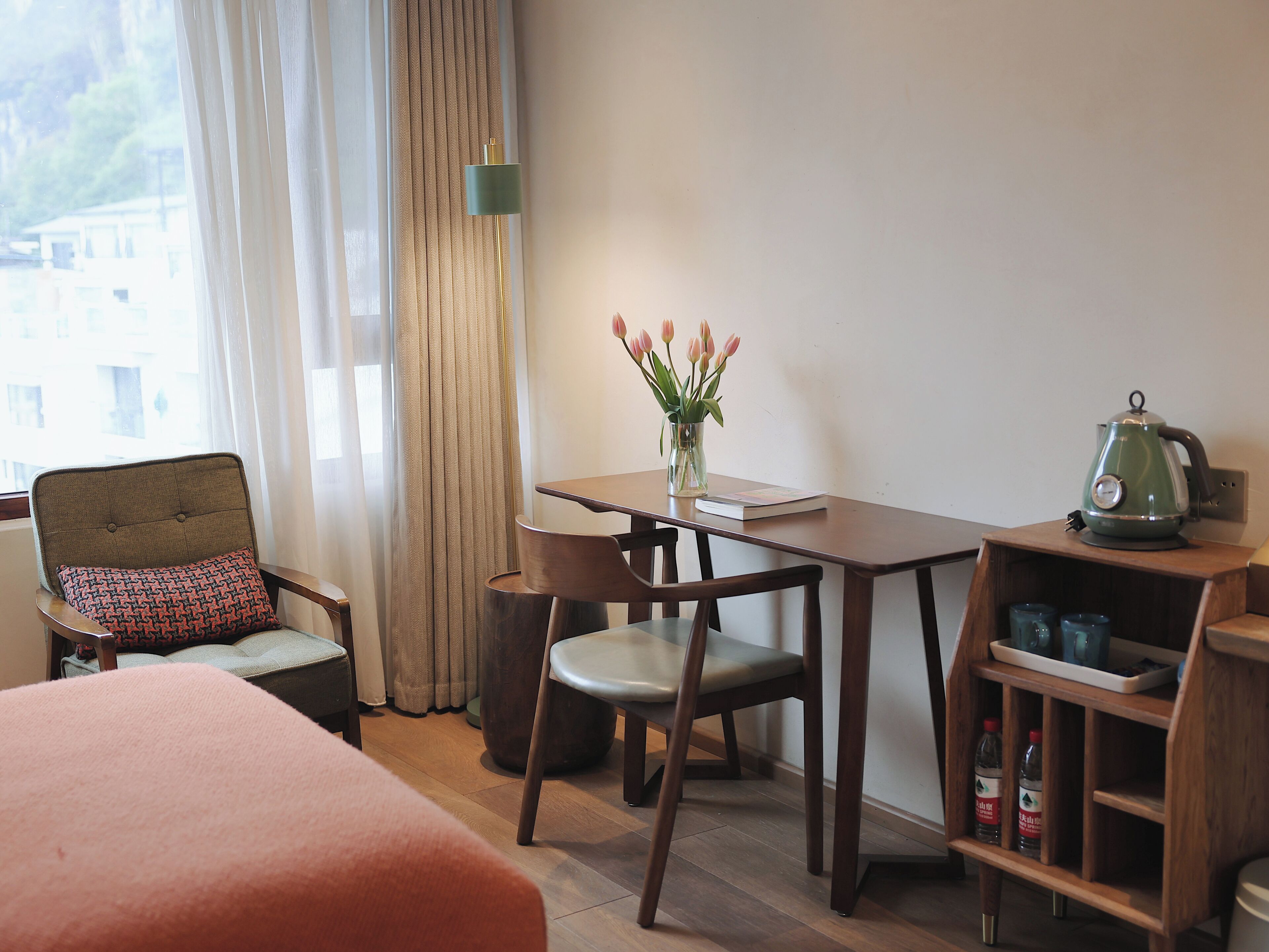 A hotel room with a bed, a table with a vase of flowers, a chair, a desk, and a kettle.