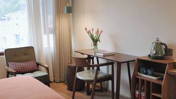 A hotel room with a bed, a table with a vase of flowers, a chair, a desk, and a kettle.