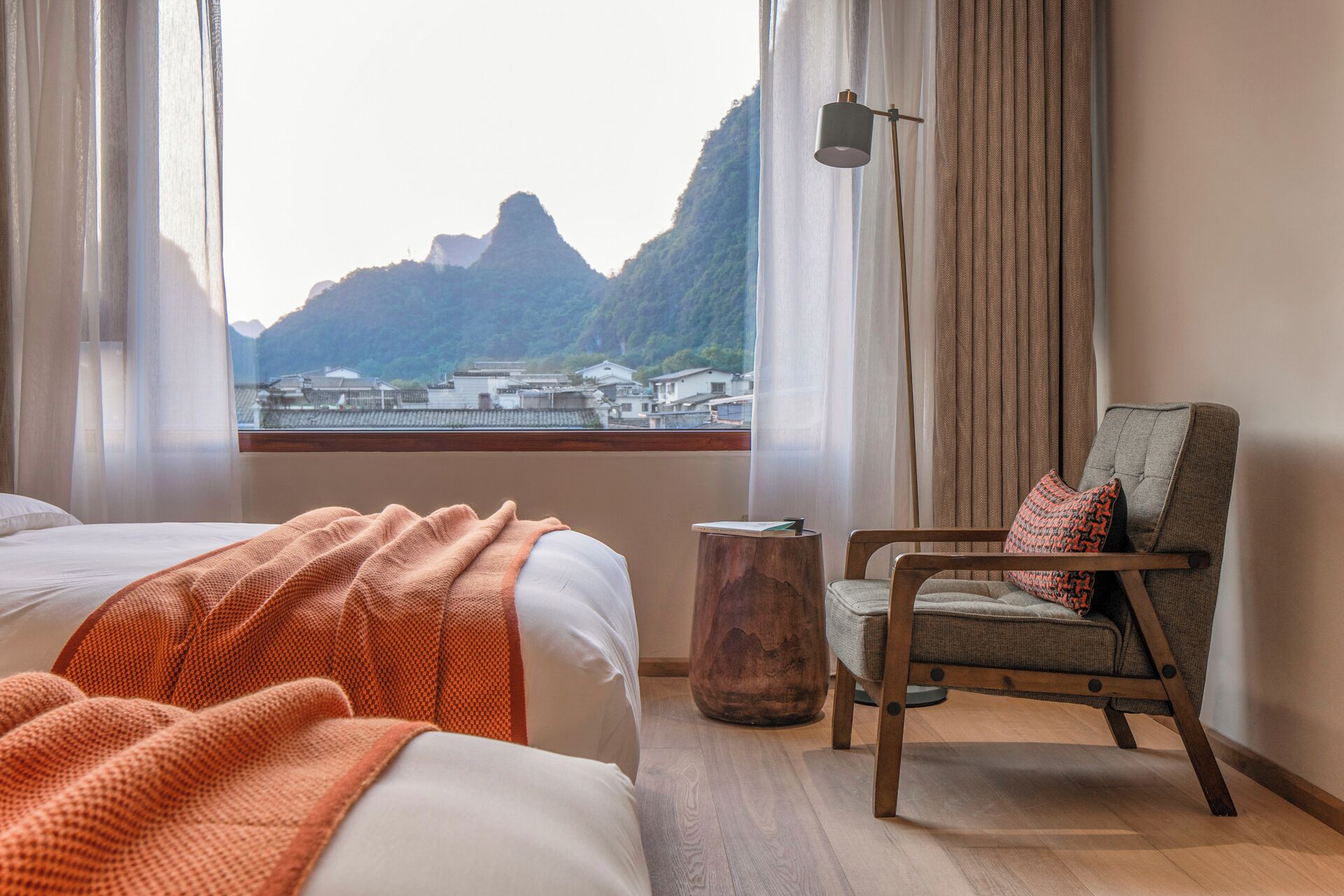 Una camera d'albergo con un letto, una sedia, uno sgabello di legno e vista su un paesaggio montuoso dalla finestra.