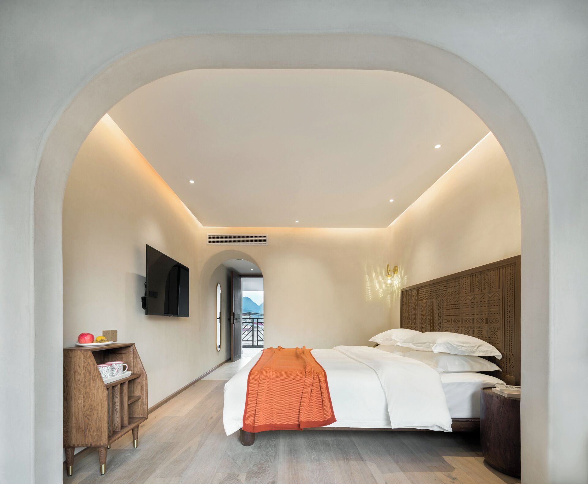 A modern hotel room with a bed, a TV mounted on the wall, a wooden side table with a cup and a book, and a view through an arched doorway.