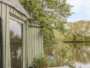A green wooden shed by a calm water body with dense foliage in the background. - Island Yurt, Beckford (Tewkesbury)