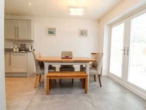 A dining area with a wooden table and chairs, a kitchen in the background, and a large window with blinds.