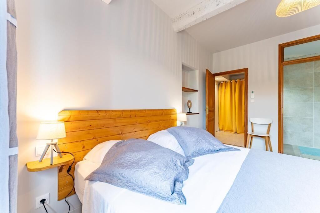Une chambre d’hôtel avec une tête de lit en bois, une table de chevet avec une lampe, un lit avec une literie bleue, une petite chaise blanche et une porte-fenêtre coulissante donnant sur une salle de bain.