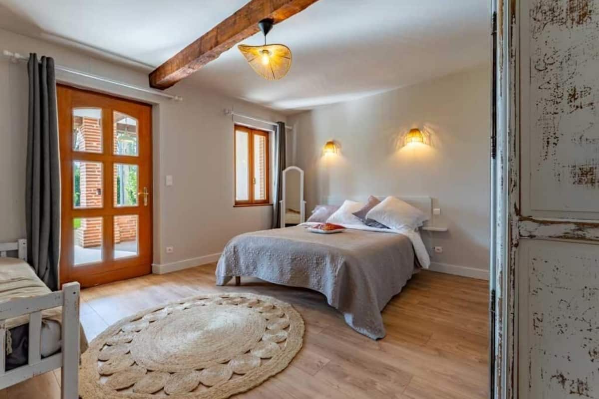 A bedroom with a bed, a wooden floor, a round rug, a door with a window, and a ceiling beam.