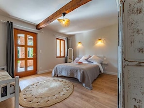 A bedroom with a bed, a wooden floor, a round rug, a door with a window, and a ceiling beam. - Ô DOMAINE (Saint-Nauphary)