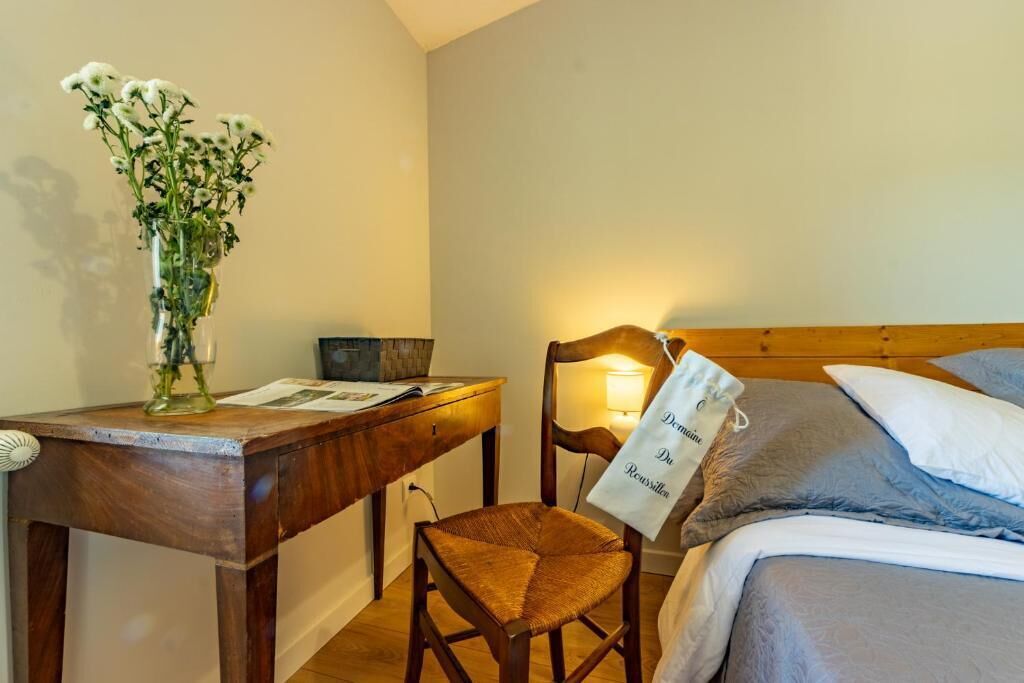 A bedroom with a wooden headboard, a bedside table with a vase of flowers, a chair, and a bag hanging on the headboard.
