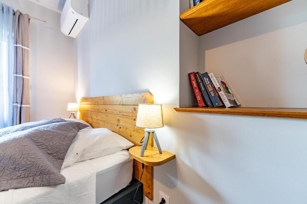 Una cama bien tendida con una manta gris, una lámpara de noche y una estantería con libros.