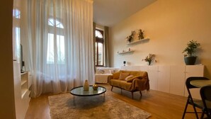 Una sala de estar con un sofá, una mesa de centro, una mesa de comedor y una silla. Hay cortinas, una planta y estantes con adornos.