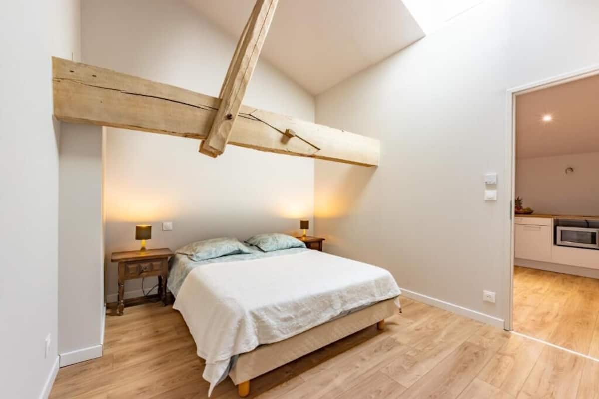 A bedroom with a wooden ceiling beam, a bed with a white bedspread, two bedside lamps, and a wooden floor.