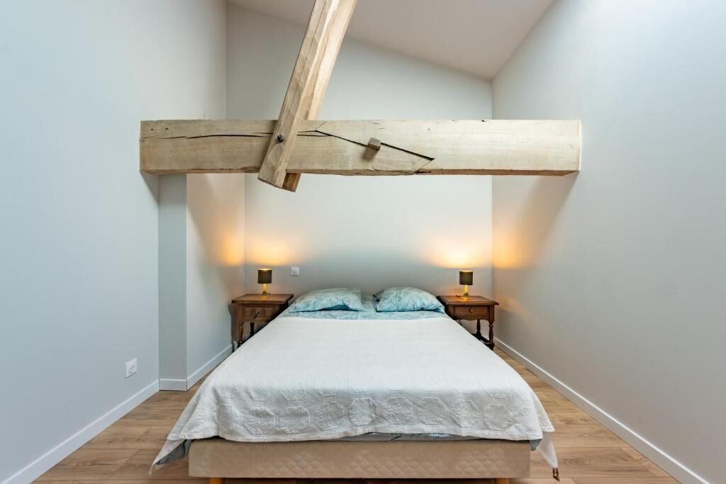 A bedroom with a wooden beam ceiling, a bed with a white bedspread, two bedside tables with lamps, and a light-colored wall.