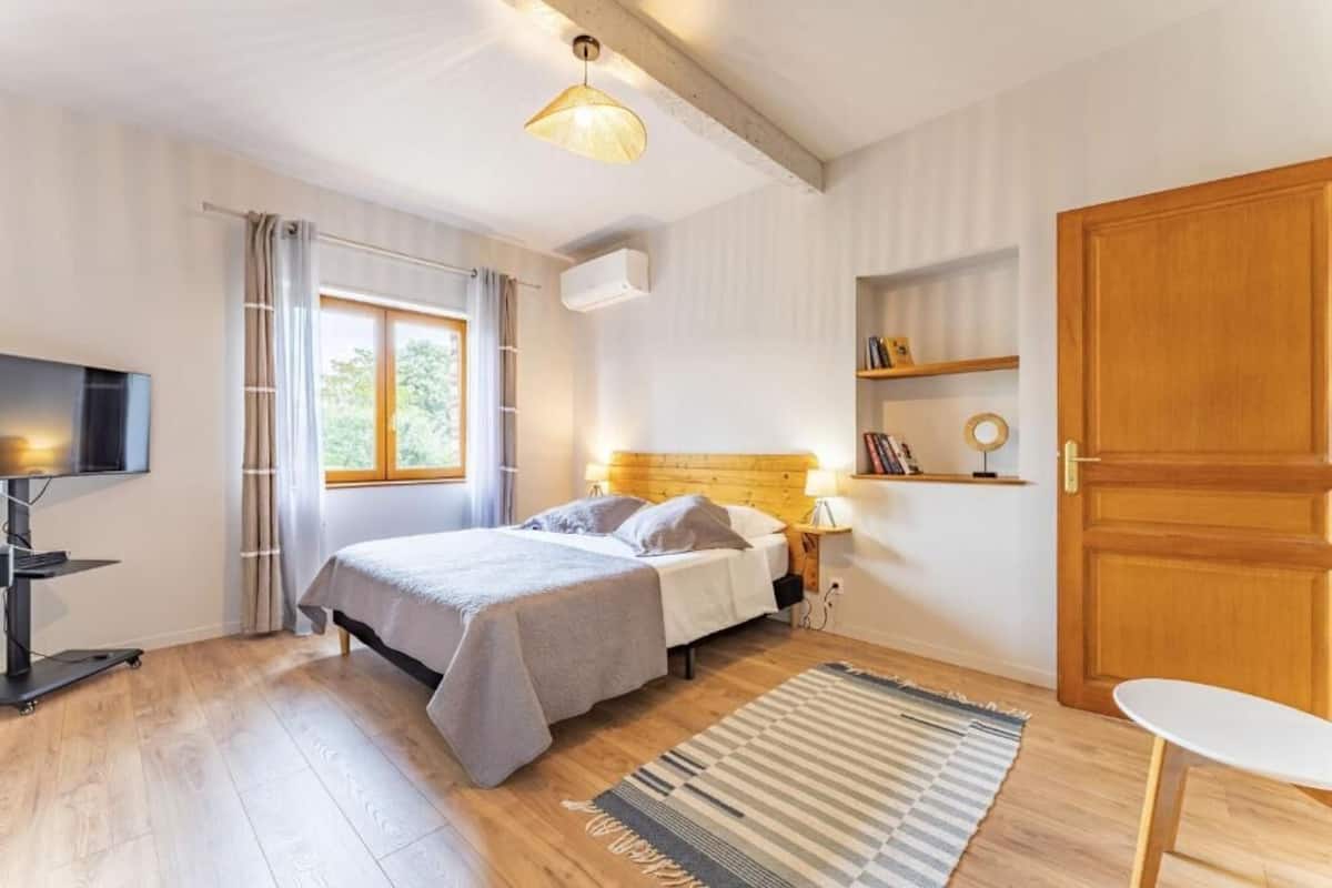 A bedroom with a bed, bedside table, lamp, TV, wooden floor, window with curtains, and a door.