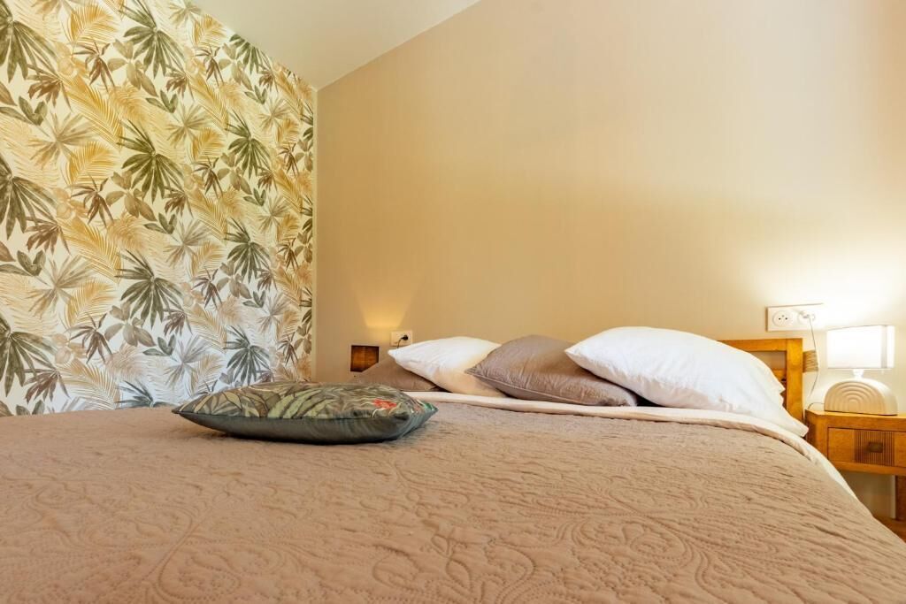 Una cama con colcha estampada, almohadas, una mesita de noche con lámpara y una pared con papel tapiz de diseño floral.