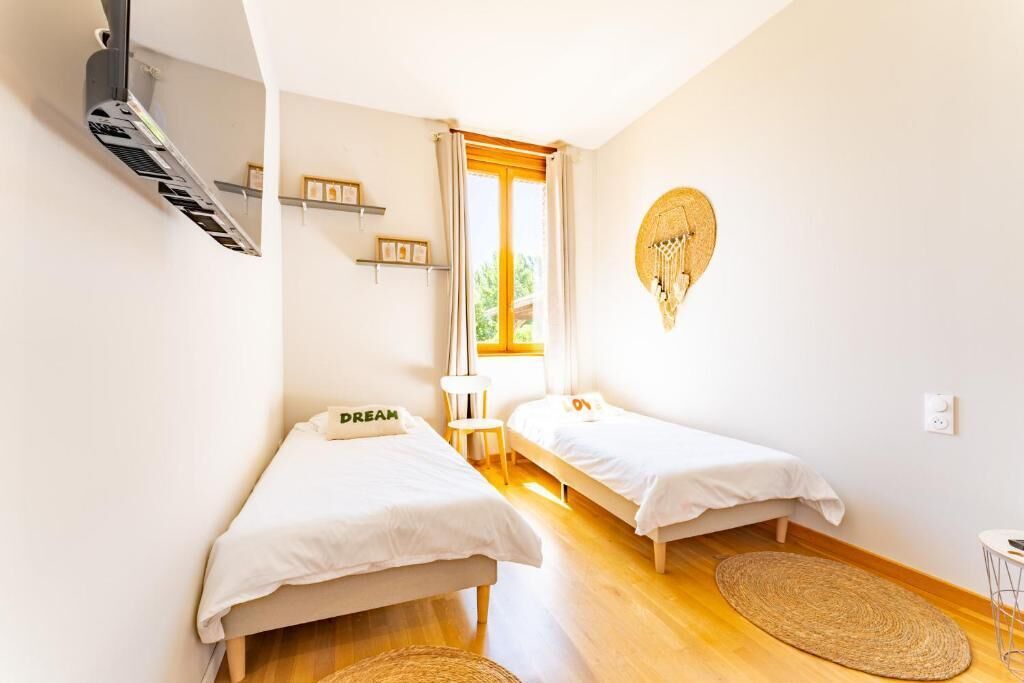Una habitación pequeña y luminosa con dos camas individuales, un ventanal con cortinas, un atrapasueños colgado en la pared y suelo de madera.