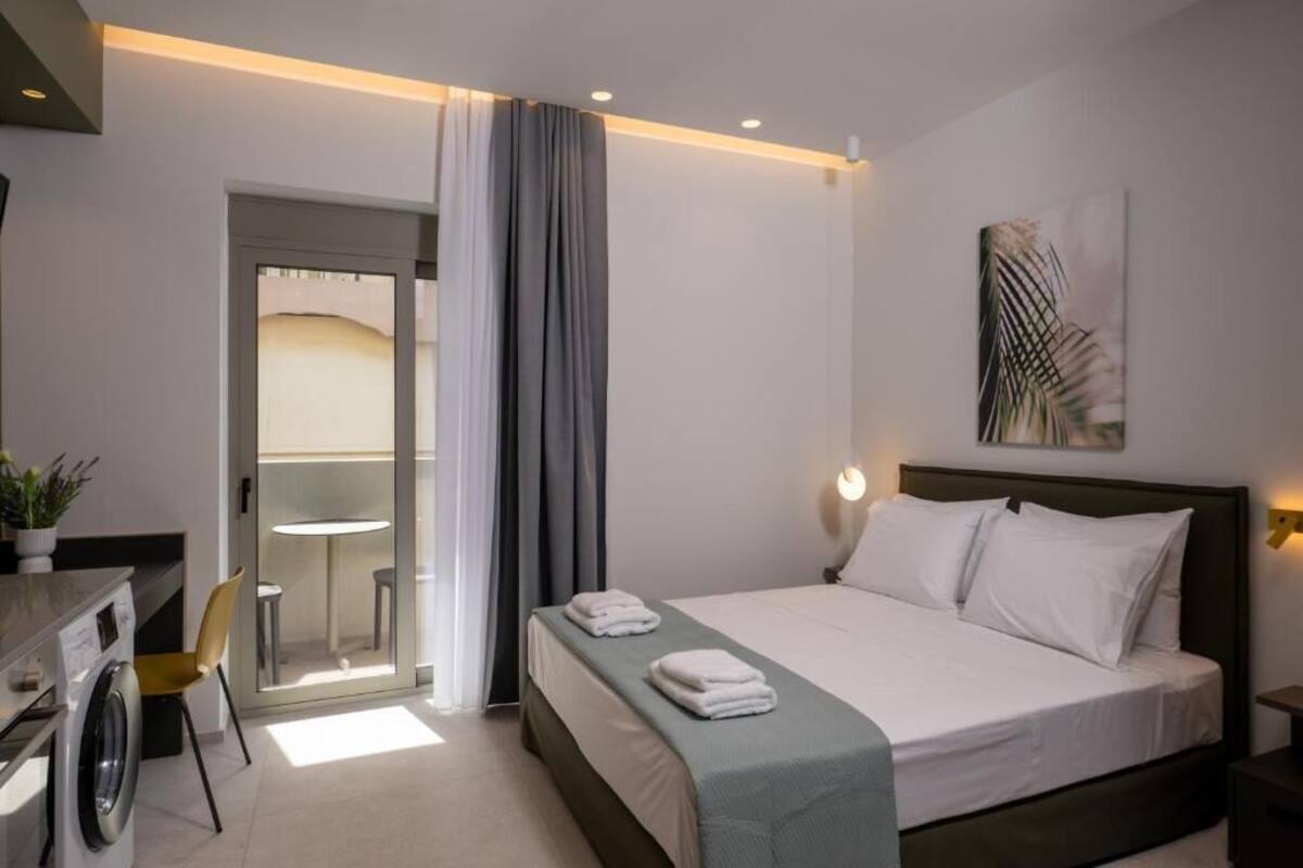 Une chambre d’hôtel avec un lit, un bureau équipé d’une machine à laver, une chaise et une vue sur l’extérieur, visible à travers une porte vitrée.
