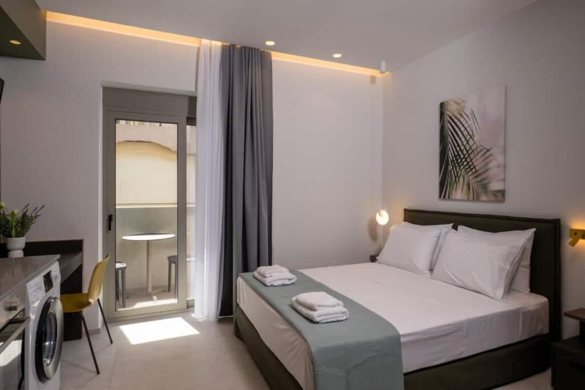A hotel room with a bed, a desk with a washing machine, a chair, and a view through a glass door.