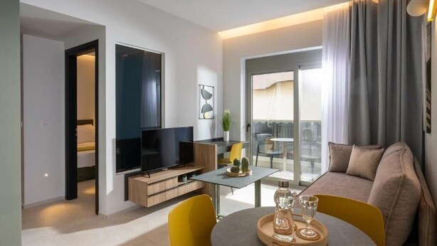 Ein modernes Wohnzimmer mit einer Couch, einem Fernseher, einem Esstisch mit gelben Stühlen und einem Balkon mit Aussicht.