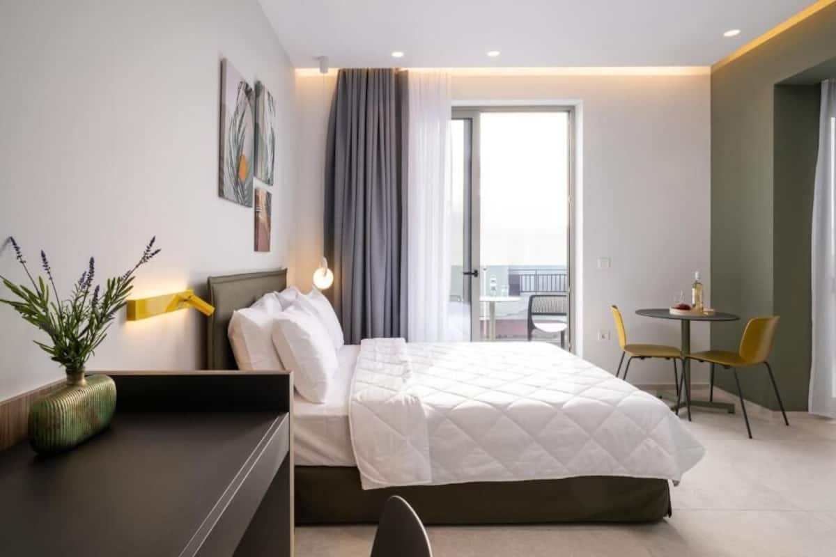 A hotel room with a bed, a desk, a chair, a small table, a plant, and a balcony with a table and chairs.