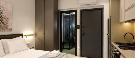 A hotel room with a bed, a bathroom with a glass door, a kitchenette, and a painting on the wall.