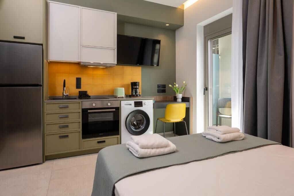 A compact hotel room with a kitchenette, a bed with two towels, a washing machine, and a TV on the wall.