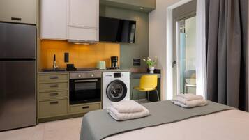 Une chambre d’hôtel compacte comprenant un coin cuisine, un lit avec deux serviettes, un lave-linge et un téléviseur fixé au mur.