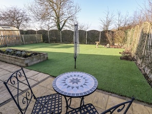 Eine Terrasse mit rundem Tisch und Stühlen, ein Gewächshaus und ein umzäunter Garten mit Rasen.