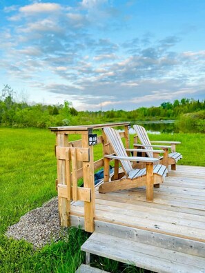 A wooden deck with Adirondack chairs and a small table, overlooking a grassy field and a body of water.