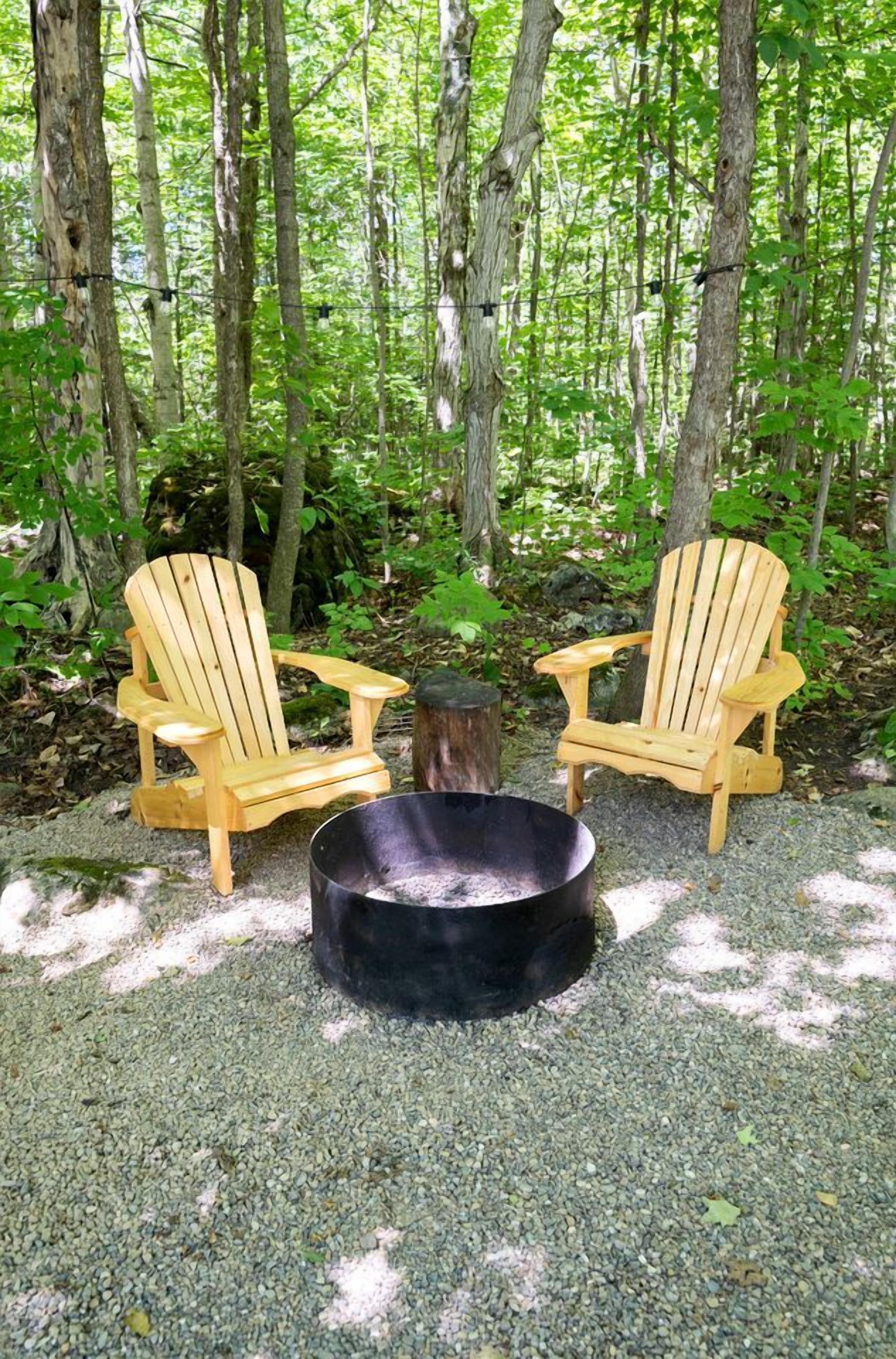 Two wooden Adirondack chairs facing each other with a black fire pit between them, set on gravel in a forested area.
