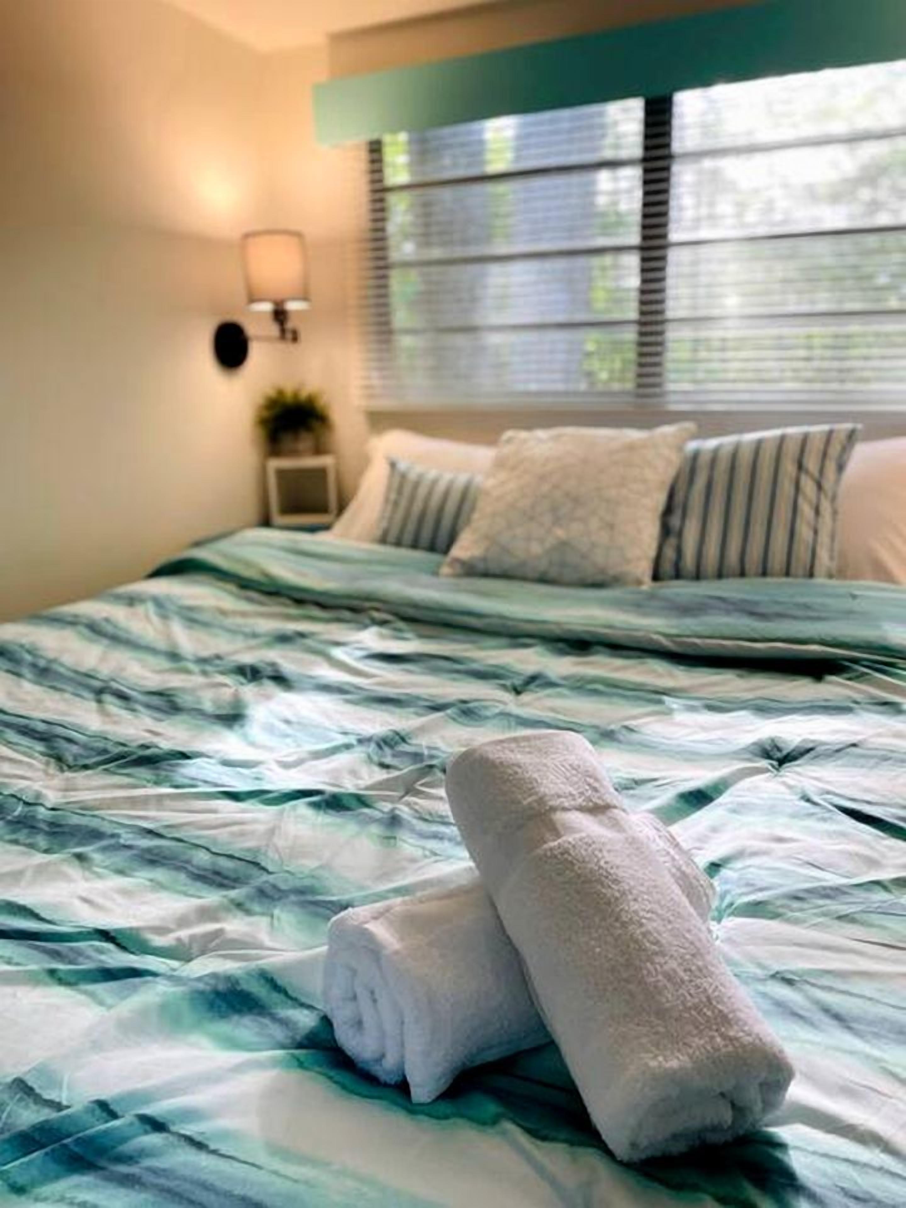 A neatly made bed with a blue and white bedspread, a pair of rolled towels, and a nightstand with a lamp.