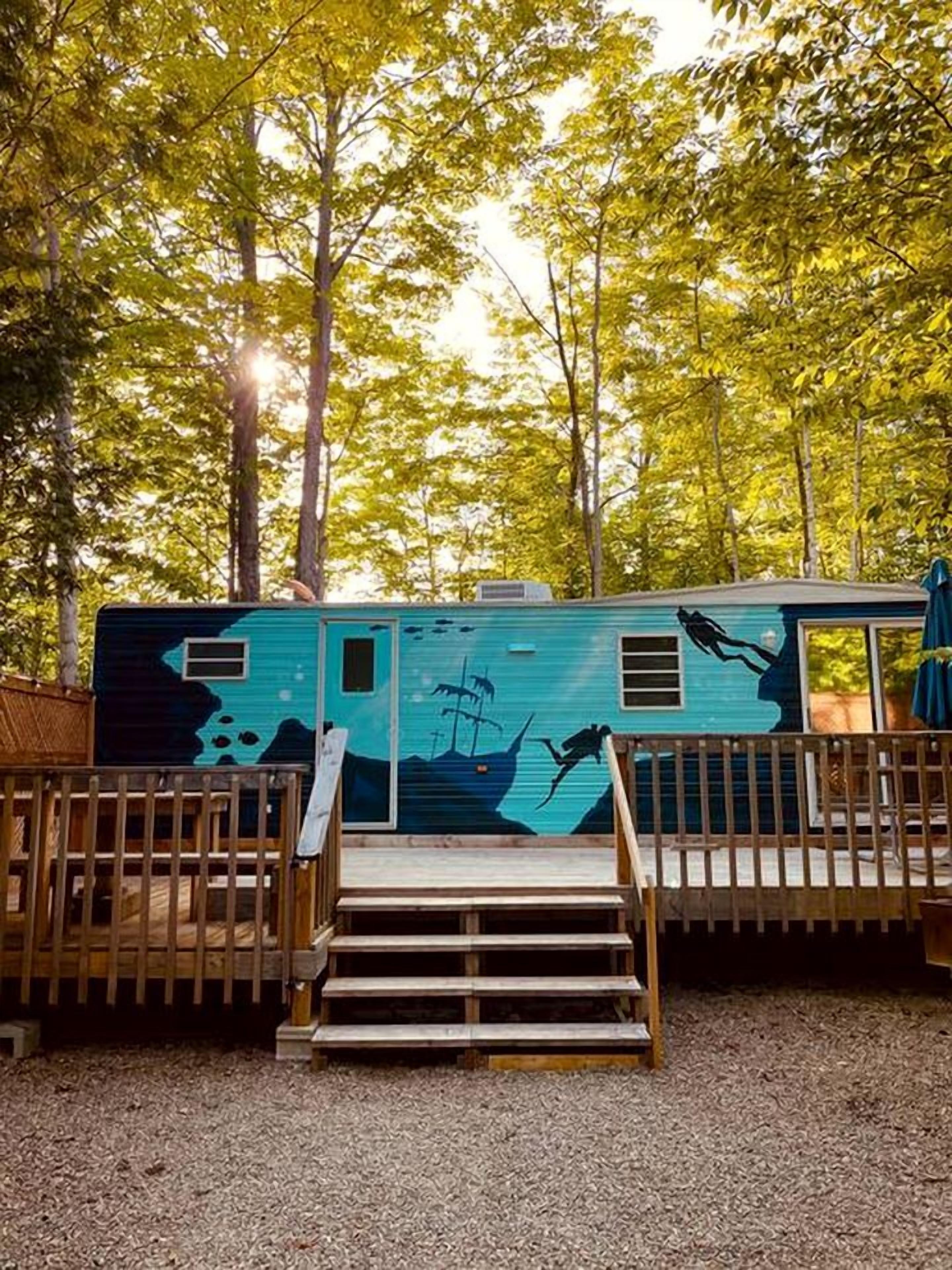 Un camping-car bleu orné d’une fresque représentant un navire et une sirène, entouré d’une forêt.