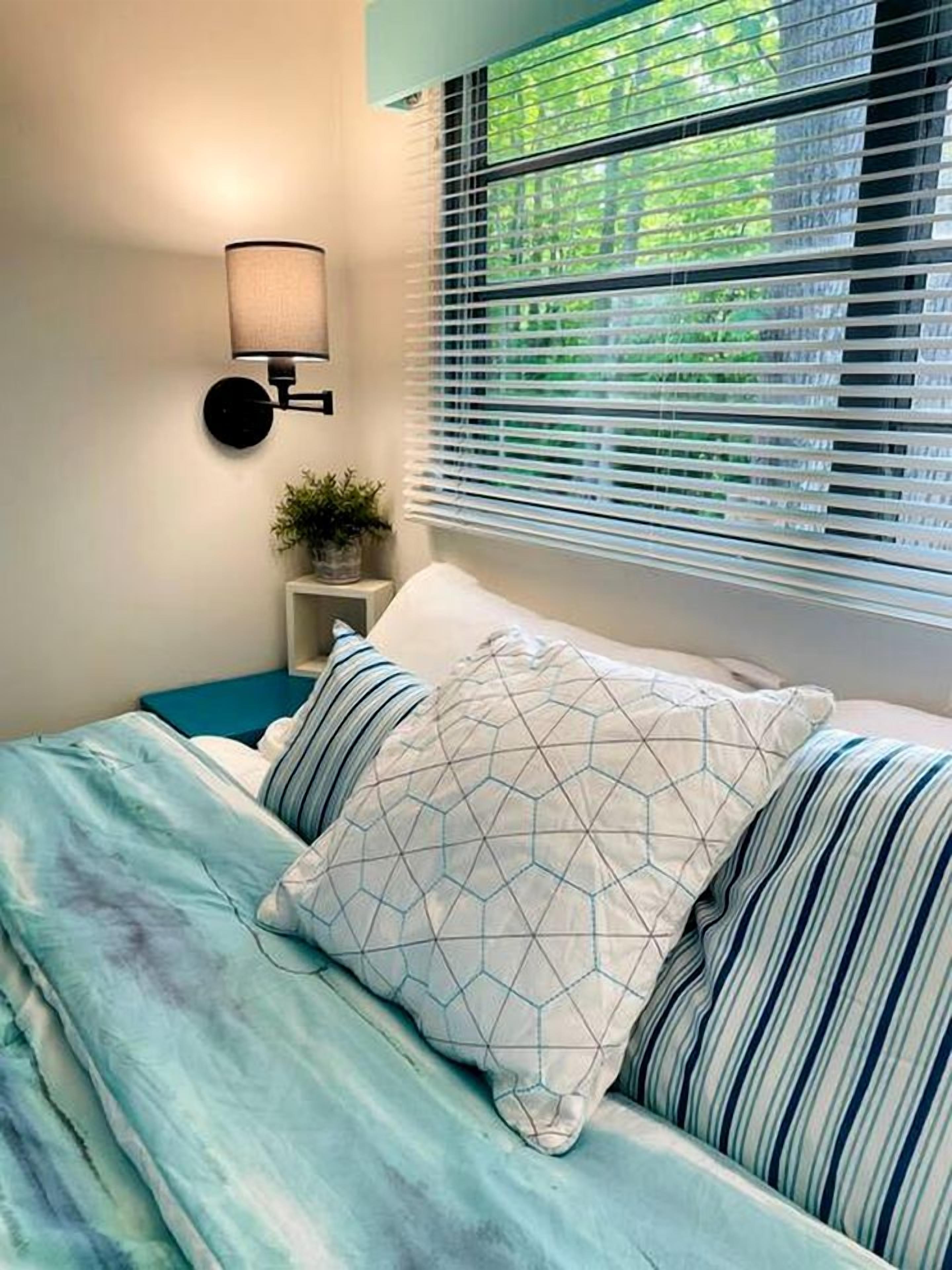 A bedroom with a bed, a wall-mounted lamp, a window with blinds, and a small bedside table with a plant.