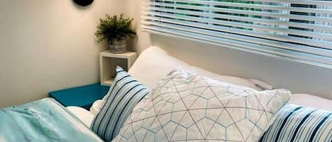 Un dormitorio con cama, lámpara de pared, ventana con persianas y una mesita de noche con una planta.