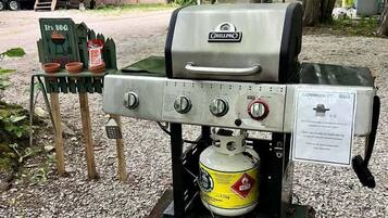 Un barbecue portatif avec une bouteille de propane, un panneau indiquant qu’il s’agit d’un espace barbecue, et un camping-car à proximité.